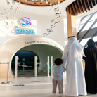 A smiling family of three stands in front of the entrance to the National Aquarium of Abu Dhabi. The parents, a husband and wife, look excited and point towards the entrance, while their young son stands between them, gazing curiously at the building. The entrance features a modern, curved design with large windows and a blue and white color scheme.