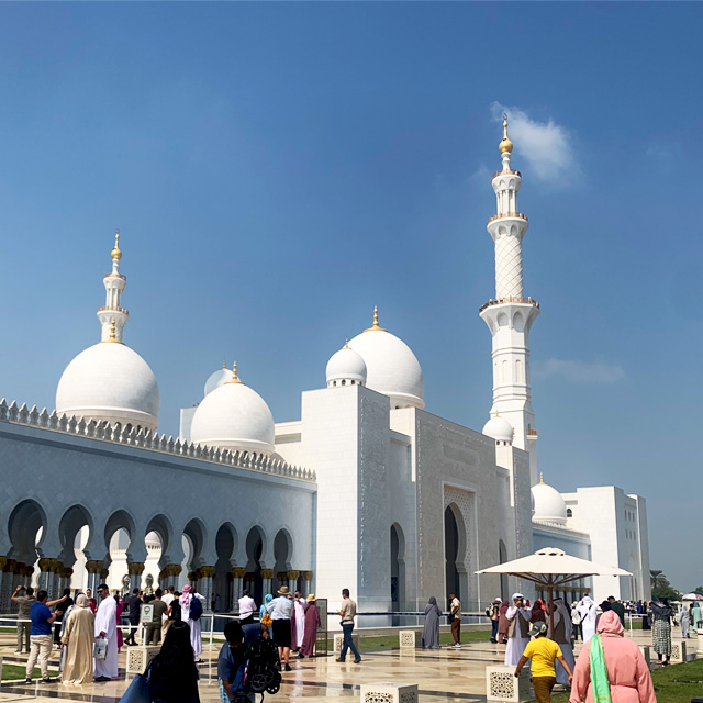 Visitors entering the Sheikh Zayed Grand Mosque in Abu Dhabi. The image features a side view of the grand entrance with its white facade, intricate details, and a towering minaret.