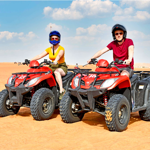 A couple riding separate single quad bikes, posing together in the vast Dubai desert against a backdrop of golden sand dunes, enjoying the Dune Rider Adventure: Off-Road ATV, Buggy Safari & Camel Ride