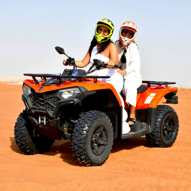Two girls wearing white outfits pose on an orange double quad bike during a Dune Rider Adventure in Dubai. They are wearing helmets for safety