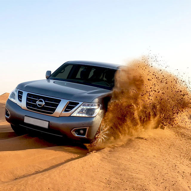 Close-up of a Nissan 4x4 vehicle tackling sand dunes on a Dubai desert safari adventure. Sand particles fly around the front left wheel as the car climbs a dune.
