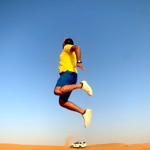 Man in a yellow shirt leaps high in the air during a Dubai desert safari adventure. A 4x4 vehicle can be seen in the background, traversing the sand dunes.