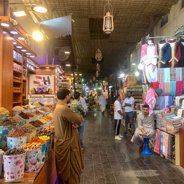 Bustling spice souk in Dubai with colorful displays of spices on the left and traditional Arabian garments on the right. Vendors showcase their wares while customers explore the vibrant marketplace.